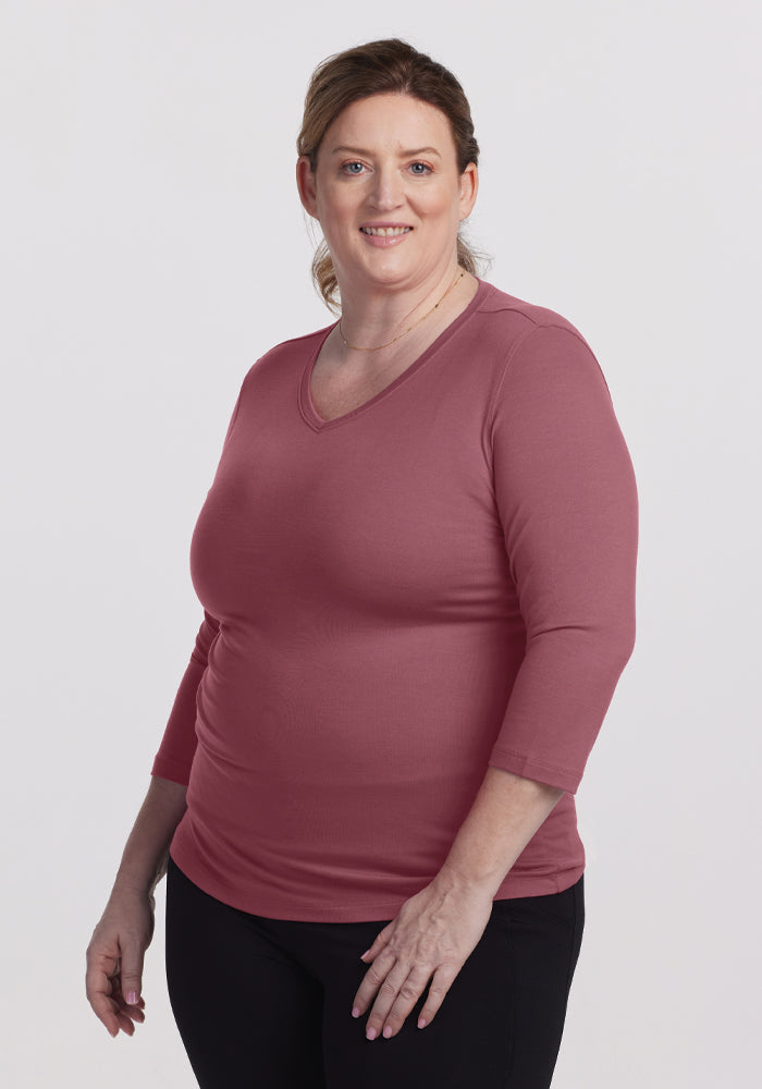 A person with medium-length hair, wearing a relaxed fit Woolx Elena V Neck in Wild Ginger and black pants, stands smiling against a plain background. 