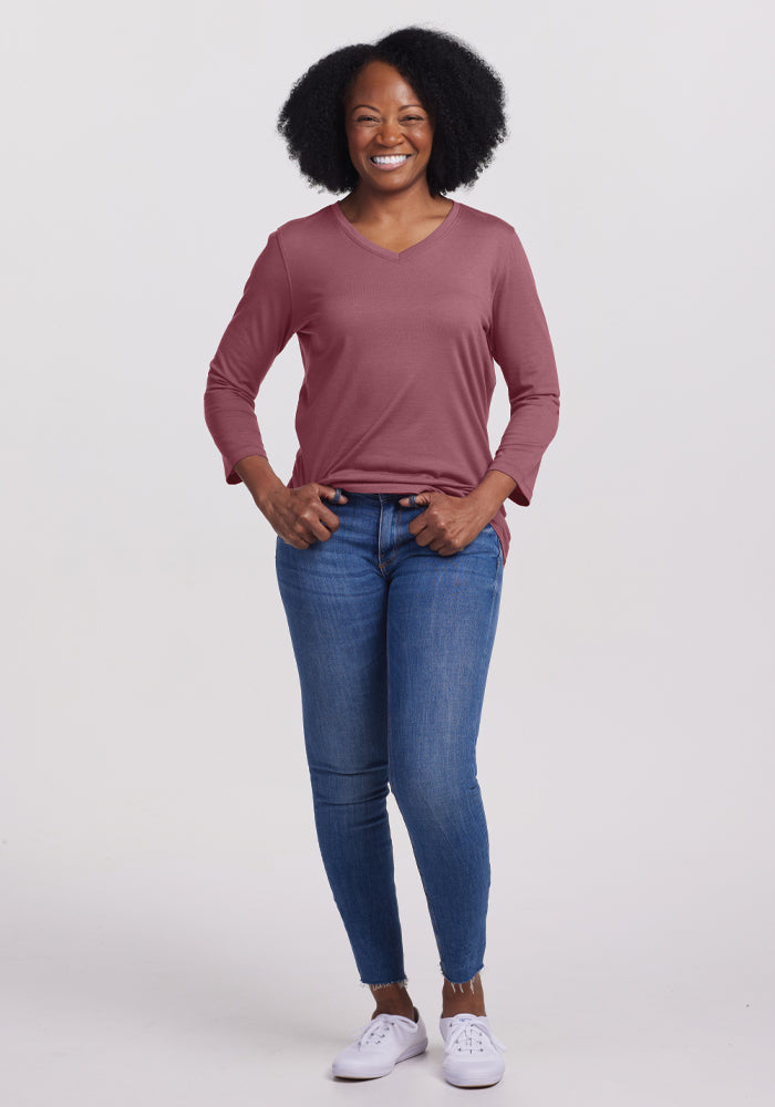 A woman with curly hair smiles while standing in a relaxed fit. She is wearing a long-sleeved, Wild Ginger Elena V-Neck Top by Woolx, blue jeans, and white sneakers. The background is plain white.