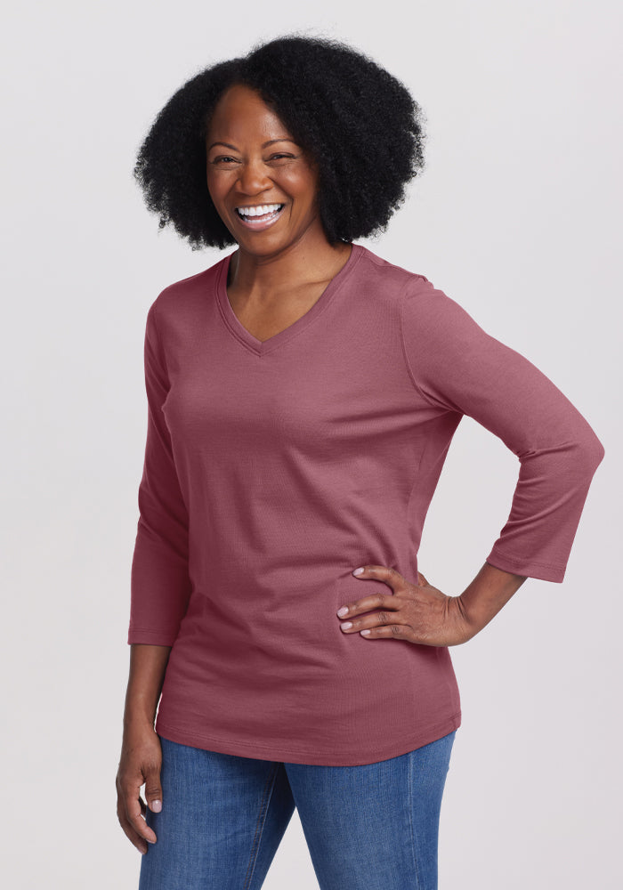 A woman with curly black hair stands with a smile, one hand on her hip. She is wearing a relaxed fit Wild Ginger Elena V Neck top from Woolx, crafted from soft Merino wool, paired with blue jeans. The background is light gray. 