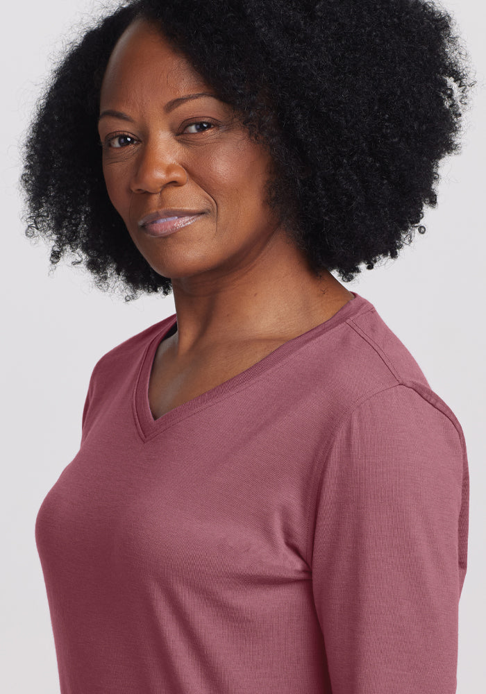 A person with curly black hair smiles slightly while wearing a relaxed fit Elena V Neck Top in Wild Ginger by Woolx. The plain, light-colored background accentuates the elegance of the Merino wool fabric.