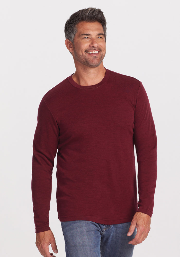 A man with short dark hair and a beard, wearing the Woolx Easton Shirt in maroon made from Australian Merino Wool and blue jeans, is smiling while looking slightly to his left. The background is plain white. 