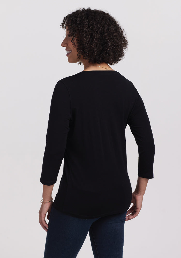 A person with curly hair faces away, dressed in a casual chic outfit featuring the Woolx Elena V Neck - Black and jeans. The background is plain and light-colored.