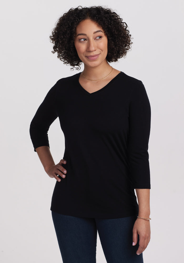 Sporting a casual chic look, a person with curly hair wears the Woolx Elena V Neck in black paired with dark jeans. Against a plain light background, they stand with one hand on their hip and glance to the side with a subtle smile. 