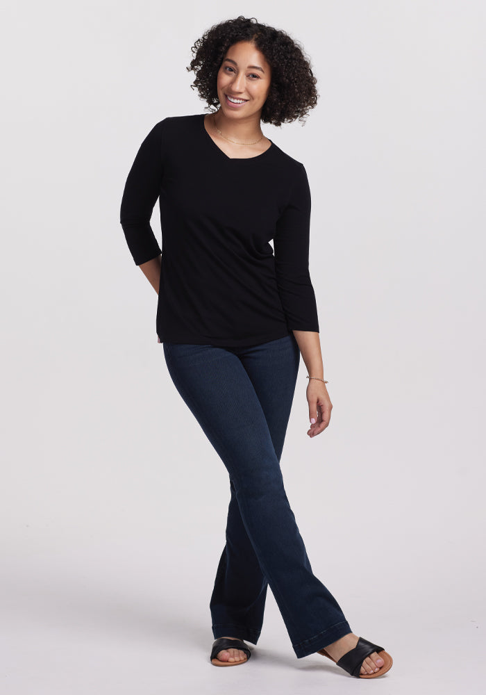 A person with curly hair poses against a plain background, wearing the chic Elena V Neck - Black from Woolx and dark blue jeans. They smile while standing with one knee slightly bent, hands relaxed near the waist.