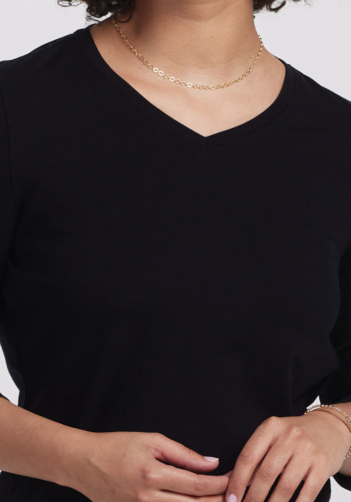 A person is shown from the shoulders down wearing a Woolx Elena V Neck - Black with a delicate gold necklace, gently touching the front of the shirt against a plain white background.