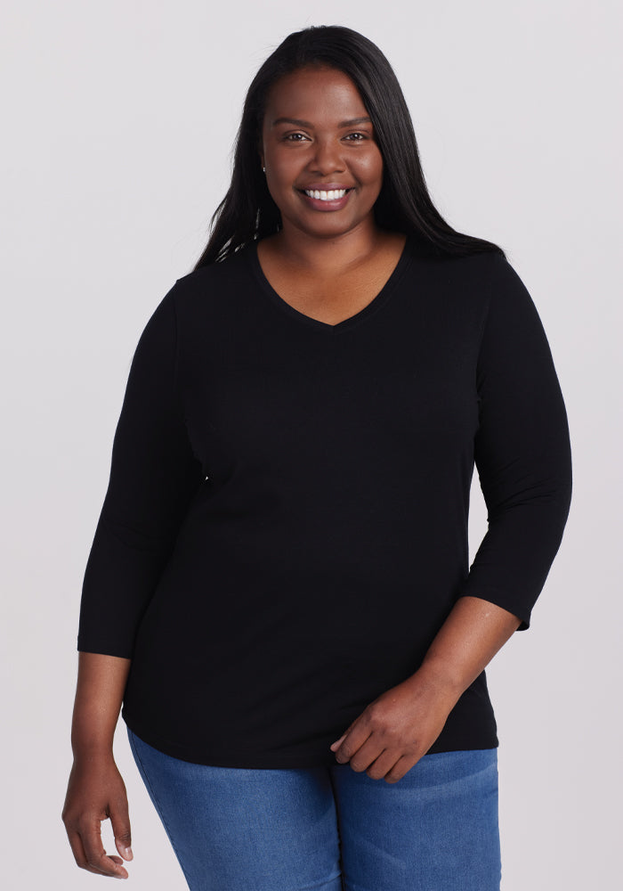 A woman smiles at the camera, showcasing her casual chic style in the Woolx Elena V Neck - Black and blue jeans, set against a plain light gray background. 