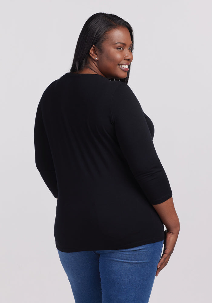 A woman with long dark hair smiles over her shoulder, exuding casual chic in the black Woolx Elena V Neck top paired with blue jeans against a plain background.