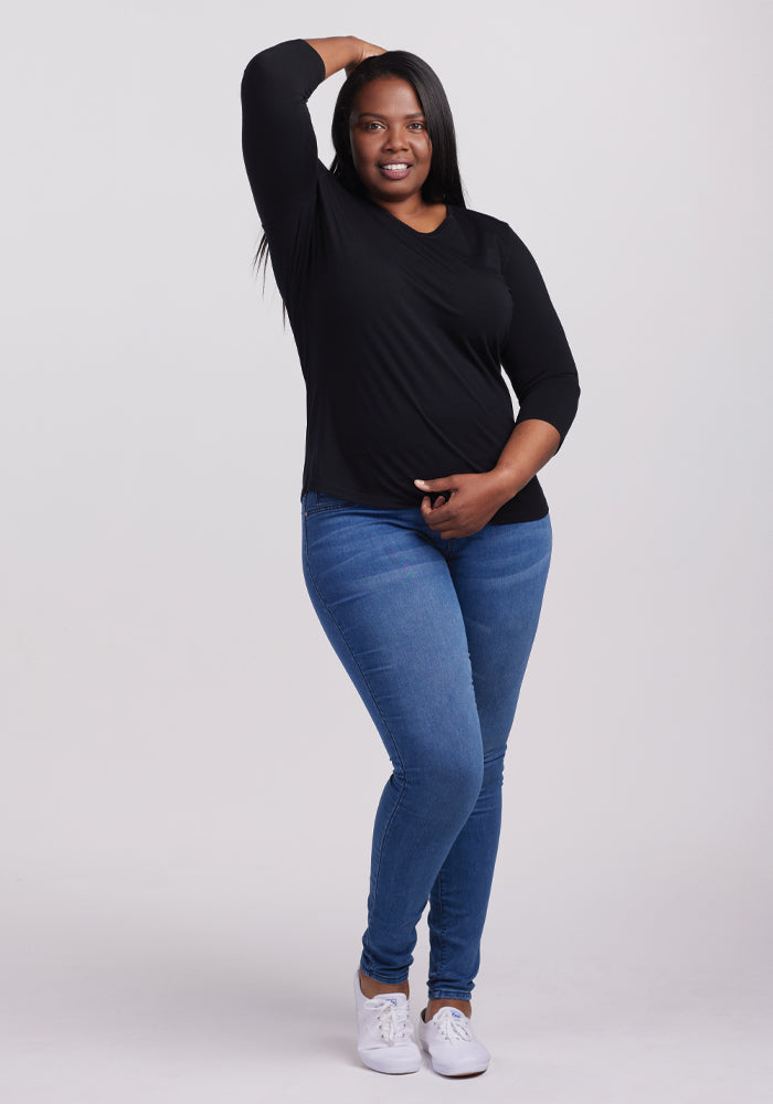 In a plain white backdrop, someone radiates casual chic in a Woolx Elena V Neck - Black, paired with blue jeans and white sneakers. They stand confidently, one hand on their hip and the other slightly elevated, smiling effortlessly.