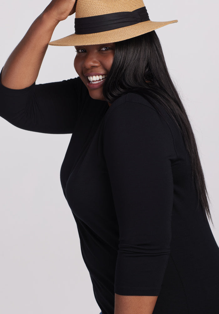 A woman with long dark hair smiles while wearing a straw hat and the Woolx Elena V Neck in black. She stands against a plain gray background, tilting the hat with one hand.