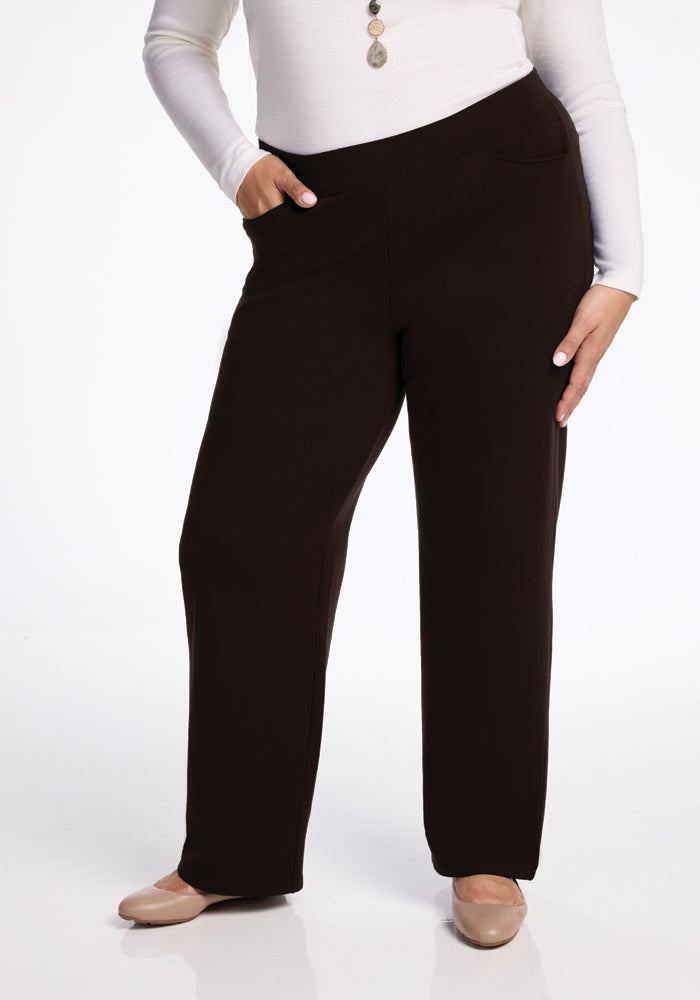 A person dressed in Woolx's Ellie Wide Leg Pants in French Roast, featuring side pockets, and a white long-sleeve top paired with a necklace stands with one hand in a pocket. They complete their look with beige flats on a reflective white floor. 