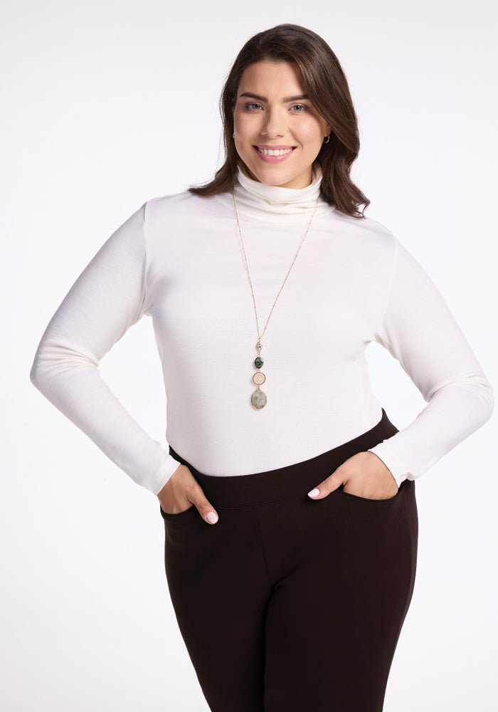 A woman with long brown hair is smiling and looking at the camera. She is wearing a white turtleneck sweater, Woolx Ellie Pants, and a long necklace with three circular pendants. Her hands are in the pockets of her warmest pants. She stands against a plain white background.