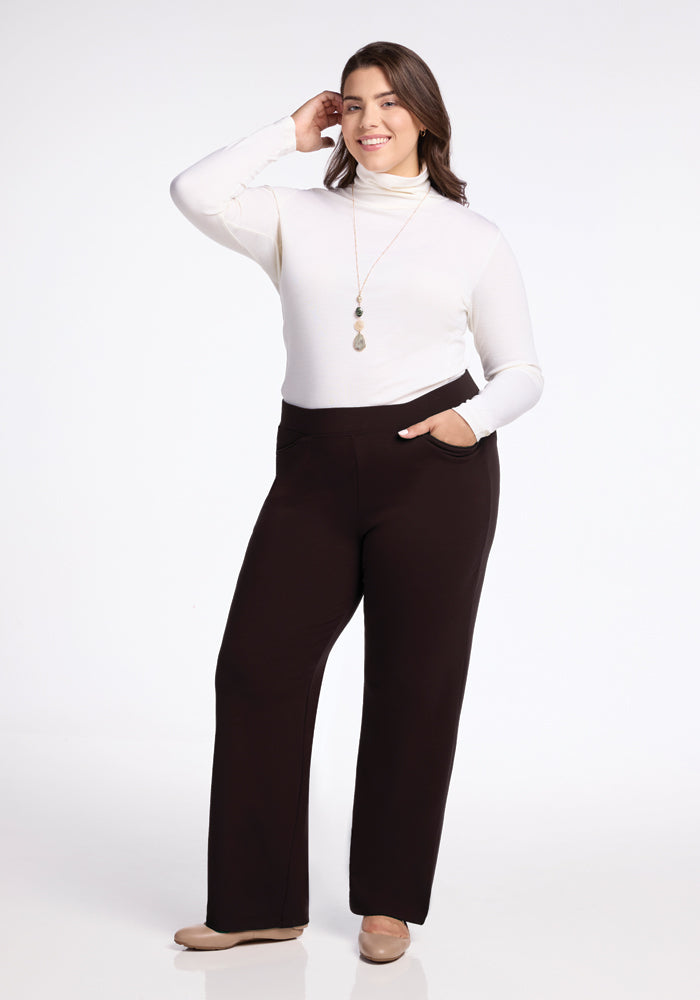 A woman with long brown hair stands against a white background, wearing a white turtleneck sweater, beige shoes, and Ellie Pants by Woolx, famous for being the warmest. She smiles gently with one hand in her pocket and the other touching her hair, adorned with a long necklace featuring a pendant.