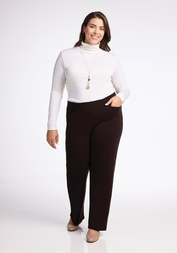 A woman is standing against a white background, wearing a white turtleneck top and Woolx Ellie Pants in black with beige heels. She has long, dark hair and is smiling. She's accessorized with a long necklace and a bracelet on her left wrist, with her left hand in her pocket.