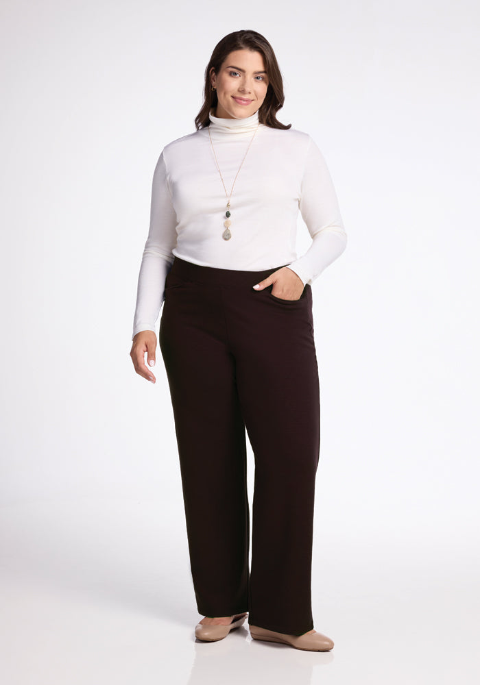 A woman poses against a plain white background, wearing a white turtleneck, Woolx Ellie Pants, and beige flats. She has one hand in her pocket and is smiling. She is accessorized with a long necklace featuring multiple pendants.