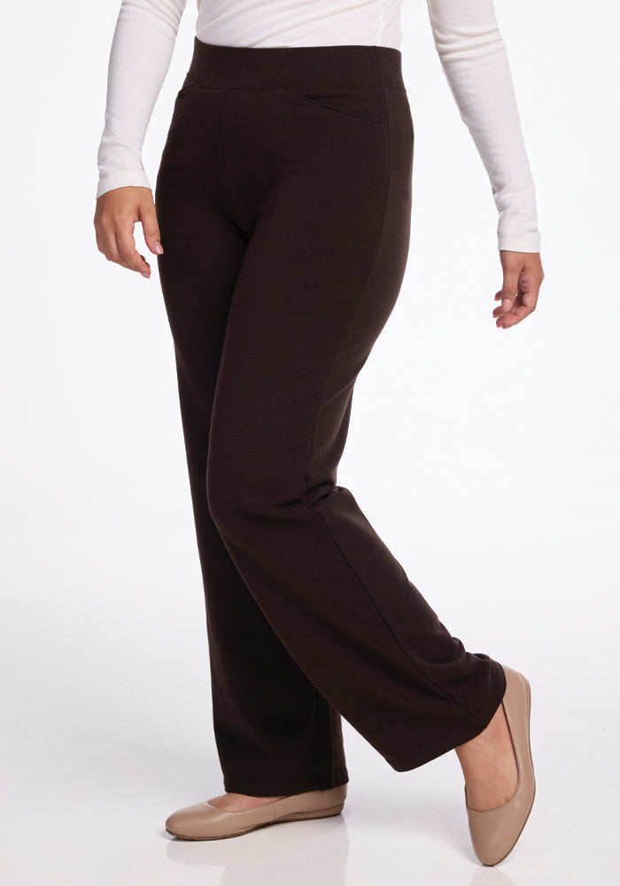 A person wearing a white long-sleeve top and high-waisted, wide-leg merino wool pants, specifically the Ellie Wide Leg Pants in French Roast by Woolx, stands against a neutral background. The outfit is completed with beige flats. The person's right foot is slightly raised, creating a relaxed posture.