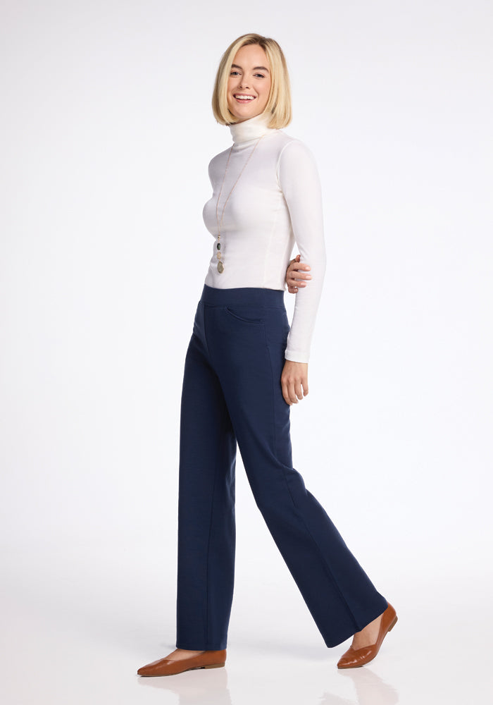 A person with short blonde hair stands smiling, wearing a white turtleneck, a long necklace, Ellie Wide Leg Pants in deep navy from Woolx, and brown shoes. The background is plain white.