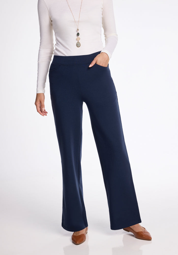 A person dressed in navy blue Ellie Pants by Woolx and a white long-sleeve top accessorized with a long necklace is standing against a plain white background. They are posing with one hand in their pocket and wearing brown pointed-toe shoes. 