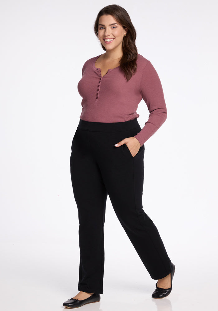 A woman stands confidently with one hand in her pocket, wearing a mauve long-sleeve top and Woolx's Emerson Straight Leg Pants Tall in black. She is smiling, her long dark hair cascading down, and completes the look with black shoes against a plain white background.