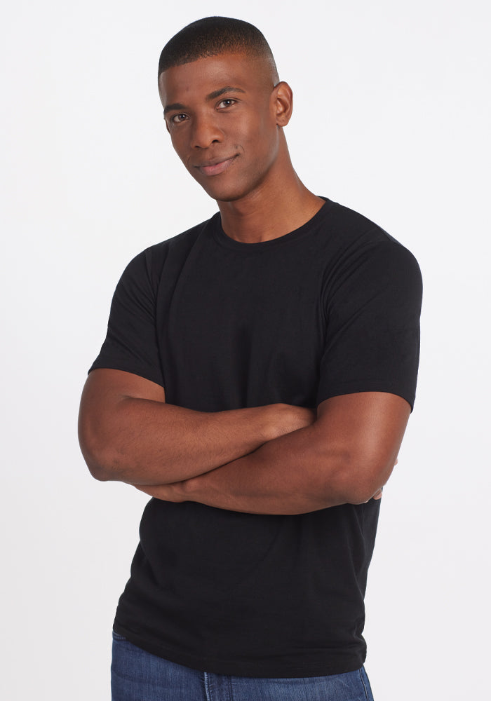 A person wearing the Woolx Endurance Tee in Black and jeans stands against a plain background, arms crossed and looking at the camera with a slight smile.