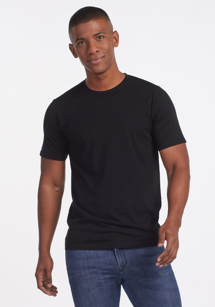 A man wearing a Woolx Endurance Tee in black and blue jeans stands smiling against a plain white background. 