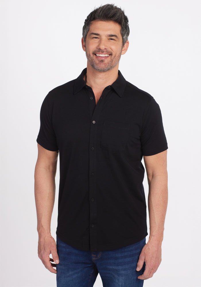 A man with short hair and a beard smiles while standing, wearing the versatile Woolx Endi Full Button Down Shirt in black made from Merino wool, paired with blue jeans against a plain white background.