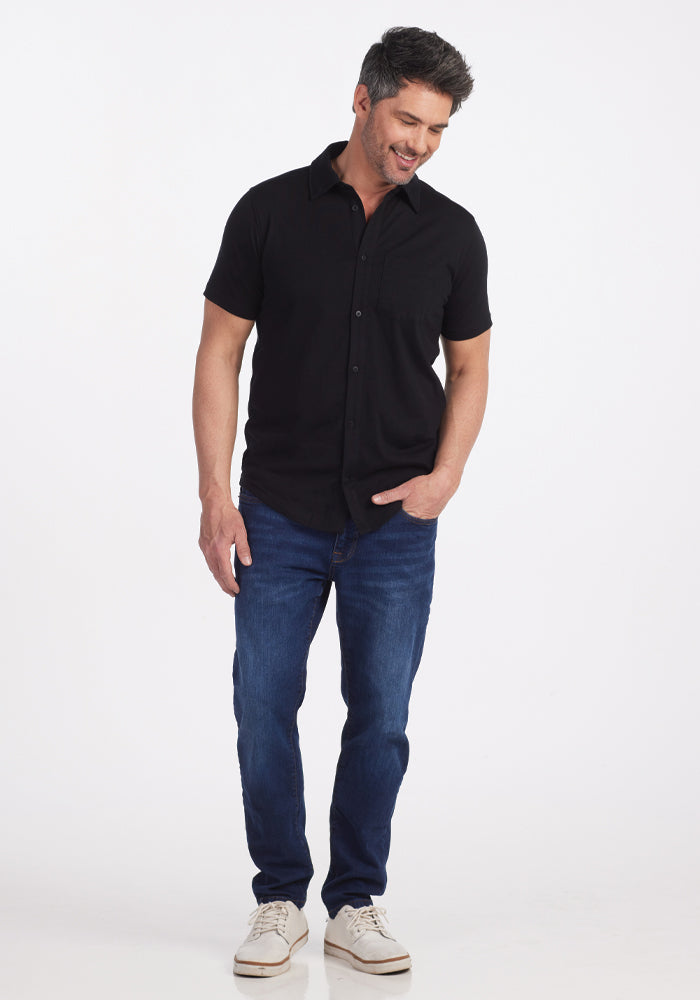 A man with short hair stands smiling, wearing a Woolx Endi Full Button Down Shirt in Black, made from versatile Merino wool. He pairs it effortlessly with blue jeans and white sneakers against a plain white background.