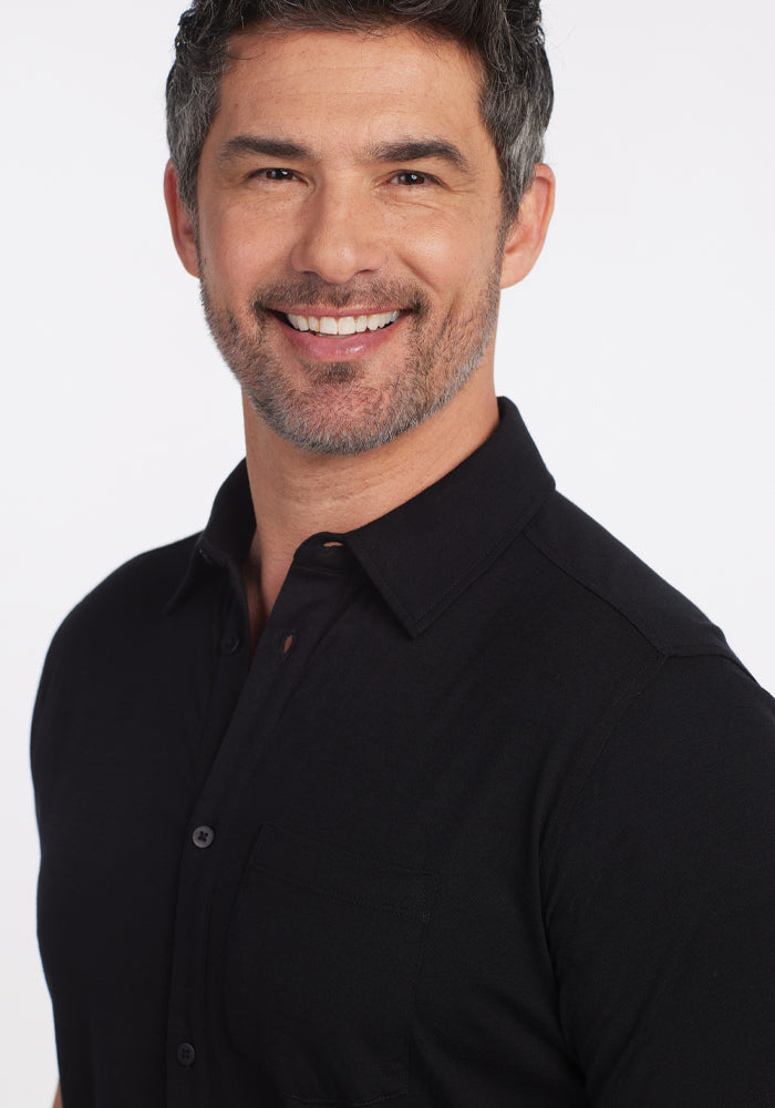 A man with salt-and-pepper hair and a beard smiles at the camera, dressed in a versatile Endi Full Button Down Shirt in black by Woolx. The background is plain white.