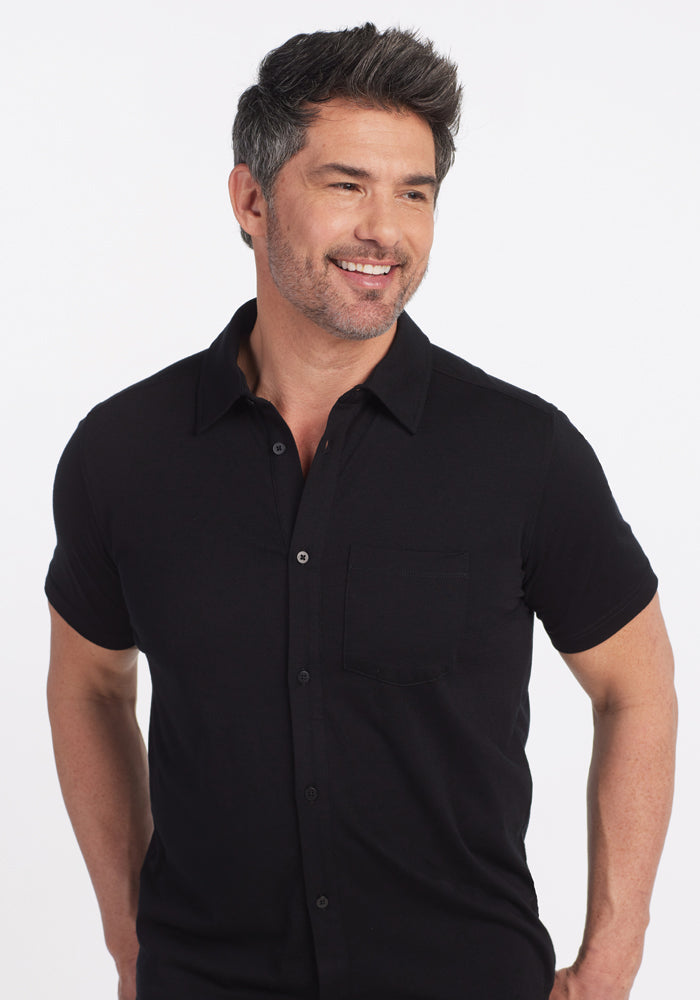 A person with short, styled hair and a beard smiles while wearing the versatile Black Endi Full Button Down Shirt by Woolx. The black button-up features short sleeves, perfectly complementing the plain white background.