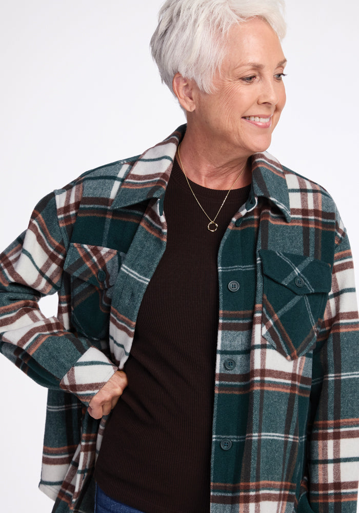 An older woman with short white hair, wearing a Woolx Sawyer Shacket in green and white plaid over a black top, smiles and looks to her right. The shacket features buttoned pockets on both sides. She appears happy and relaxed.