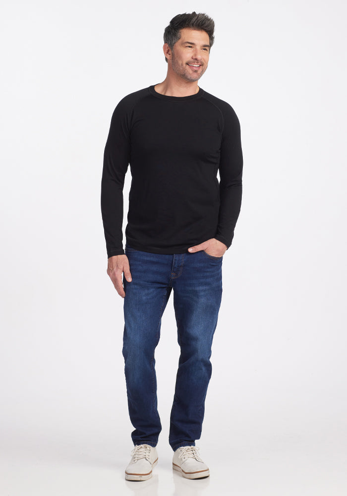 A man wearing the Woolx Essential Tee in black, paired with blue jeans and white sneakers, stands smiling with one hand in his pocket against a plain white background.