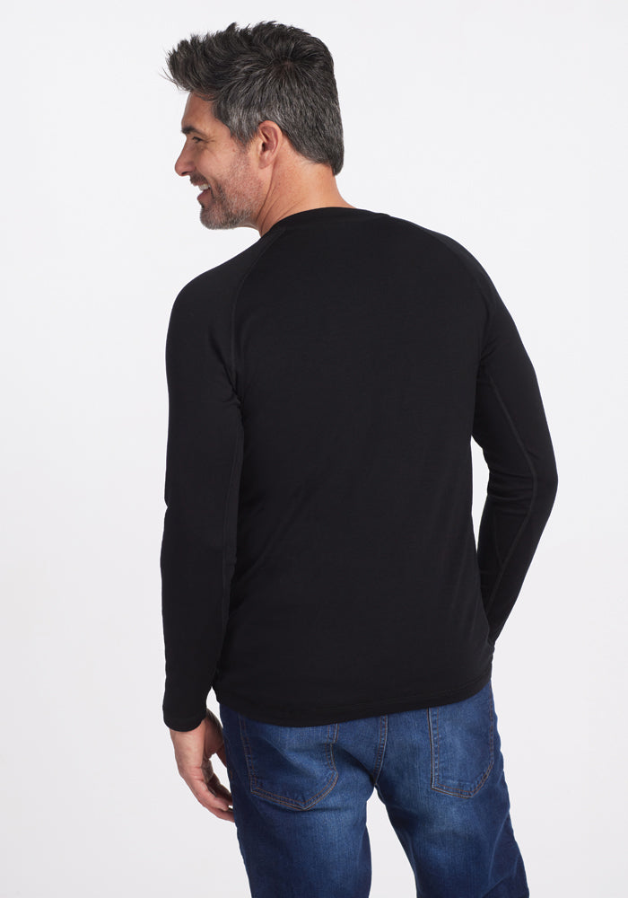 A man with gray hair and a beard is smiling and looking to the side against a plain white background, wearing the Woolx Essential Tee in black along with blue jeans.