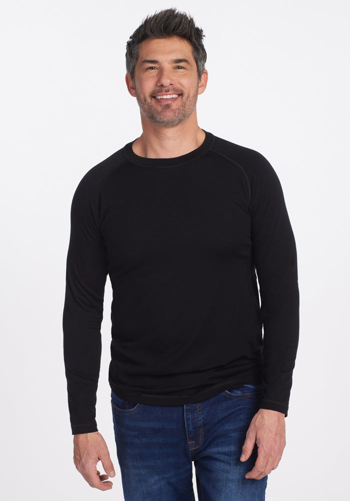 A man with short, dark hair and a beard smiles while wearing a black long-sleeve Essential Tee from Woolx and blue jeans against a plain white background. 