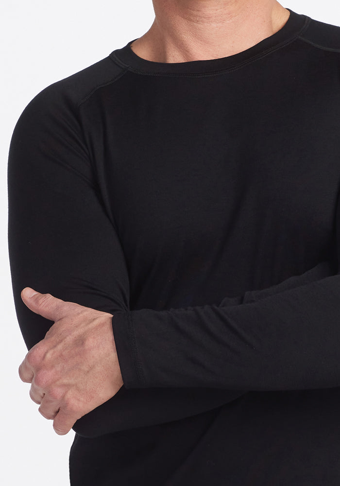 A person wearing a black Woolx Essential Tee stands with arms crossed against a white background, showcasing only the upper torso and arms.