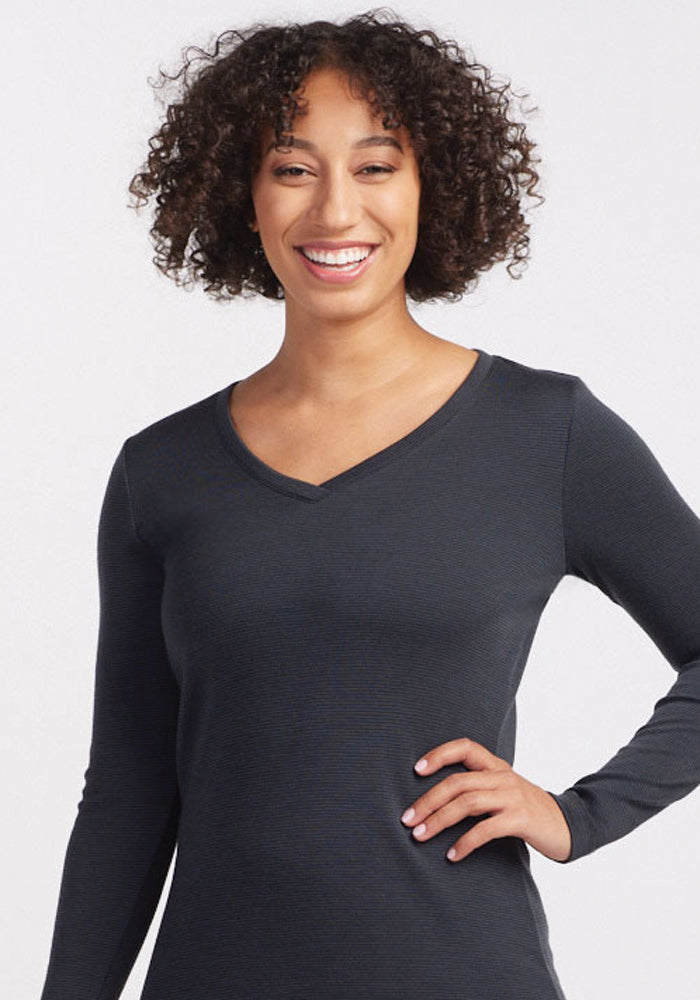 A woman with curly hair smiles at the camera, wearing the Eva Tunic in Carbon Stripe from Woolx. With one hand on her hip, she stands confidently against a plain white background.