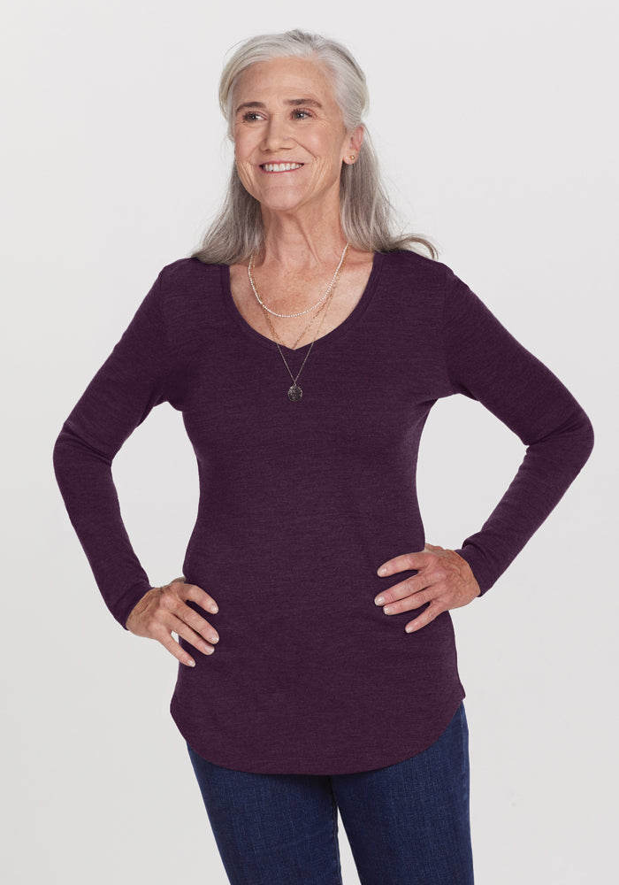 A smiling older woman with long gray hair wears the Woolx Eva Tunic in Deep Plum and dark jeans. She poses with her hands on her hips against a plain white background. She is also wearing two layered necklaces. The cozy Australian Merino Wool tunic adds a touch of elegance to her street style.