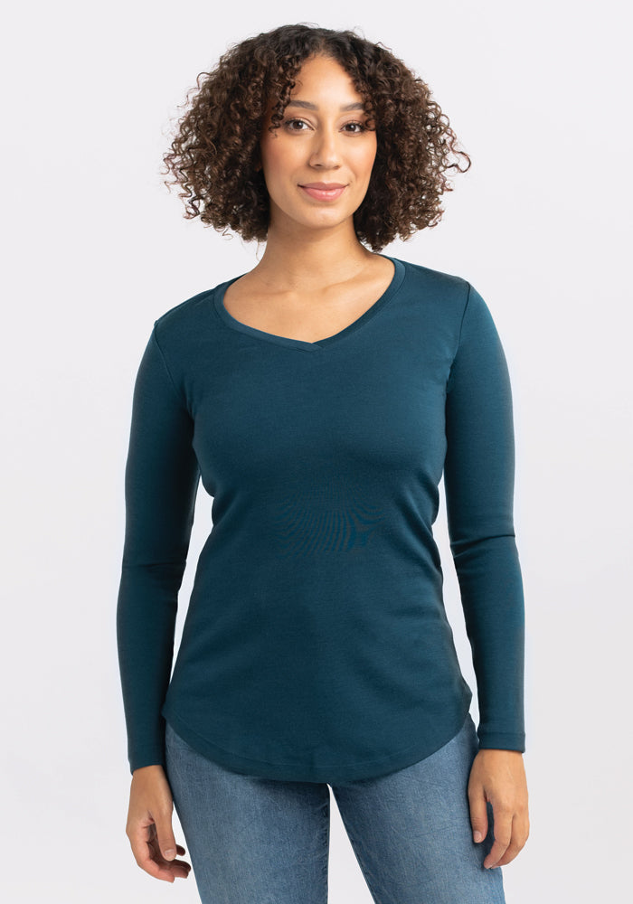 A person with curly hair is standing and facing the camera, wearing the Woolx Eva Tunic in teal and blue jeans. The background is plain white. 