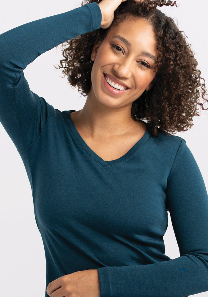 A smiling woman with curly hair poses with one arm raised and her hand resting on her head. She is wearing a long-sleeved, dark teal Eva Tunic from Woolx. The background is plain and light-colored.