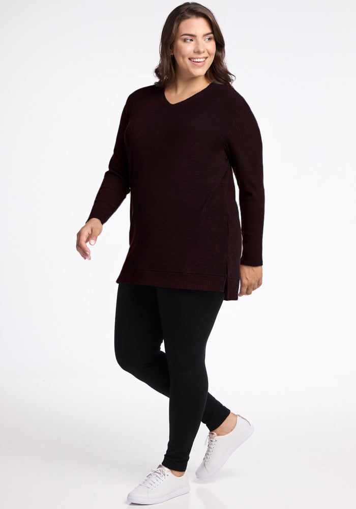 A person with shoulder-length hair is smiling and walking. They are wearing the cozy and warm Evie Ribbed Tunic from Woolx, paired with black leggings and white sneakers. The background is plain white. 