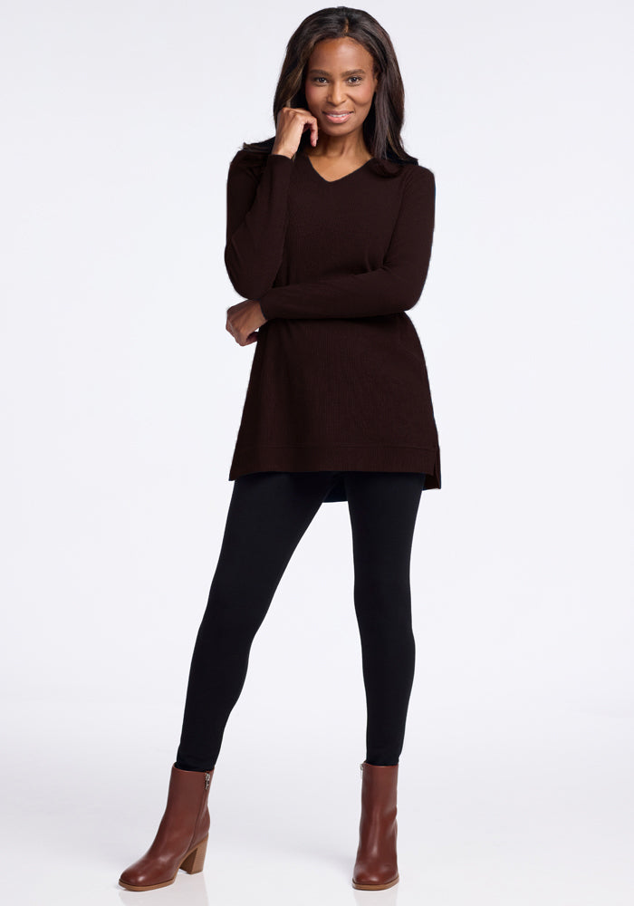 A person stands confidently against a plain background, wearing the cozy and warm Evie Ribbed Tunic from Woolx, which features a flattering V-neck fit. They are paired with black leggings and brown ankle boots. Their long hair cascades as they smile slightly, with one hand resting on their cheek. 