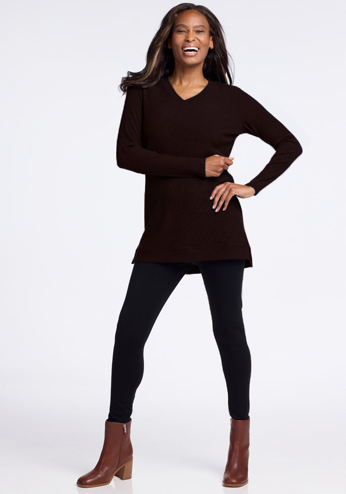 A woman stands smiling with her hand on her hip, showcasing a flattering V-neck fit. She is wearing the Woolx Evie Ribbed Tunic, a long-sleeve black sweater made of cozy and warm Merino wool, paired with black leggings and brown ankle boots with heels. The background is plain white.