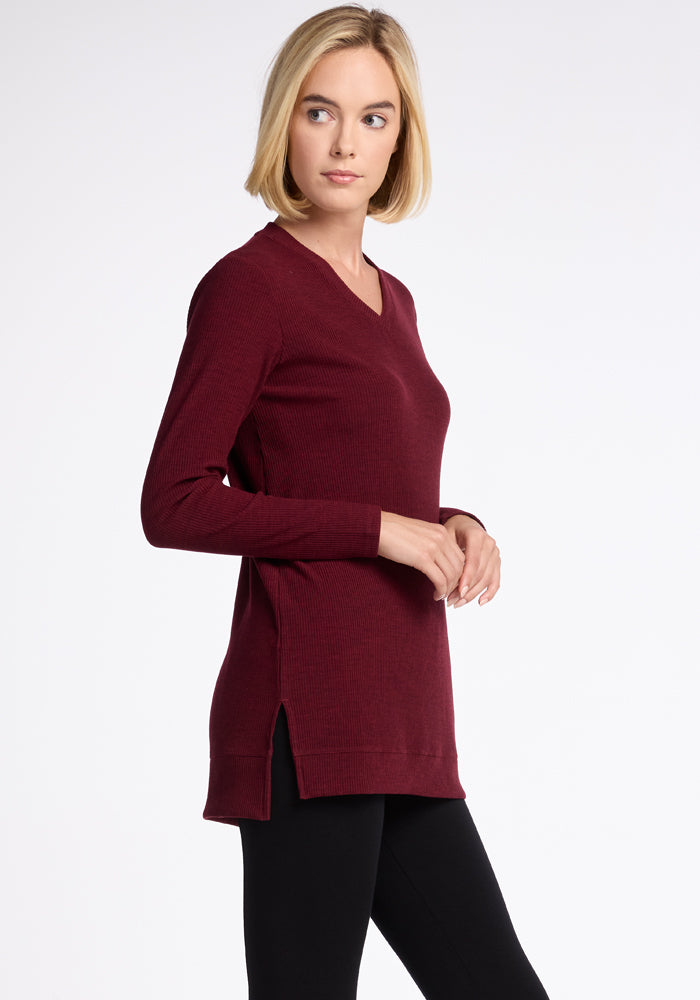 A woman with blonde hair is wearing the Woolx Evie Tunic in Cranberry Melange paired with black leggings. The cozy long-sleeved V-neck sweater drapes elegantly as she stands sideways with her hands together, looking to her left against a white background. 