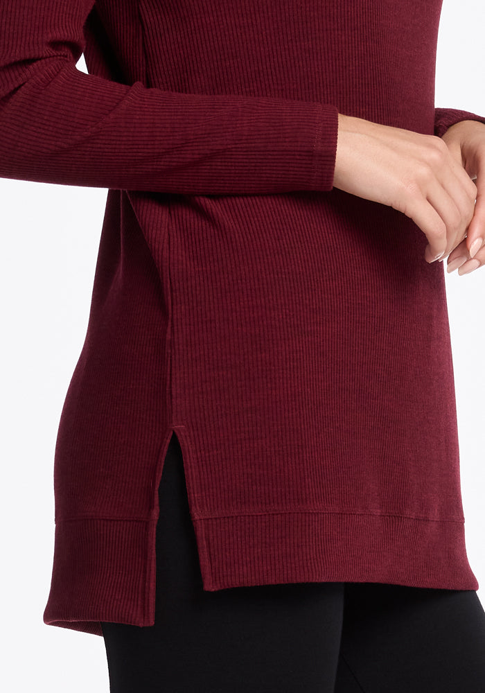 A person wearing the Woolx Evie Tunic in Cranberry Melange, a long-sleeved, ribbed sweater with a side slit, is visible from mid-chest to thigh, standing in a relaxed pose with one hand resting near their waist. The background is plain white.