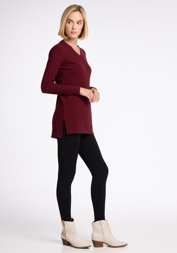 A woman with short blonde hair is wearing the Evie Tunic - Cranberry Melange by Woolx, along with black leggings, and cream ankle boots. She stands in front of a plain white background, posed in profile with her right hand resting on her abdomen and her left hand relaxed by her side. 