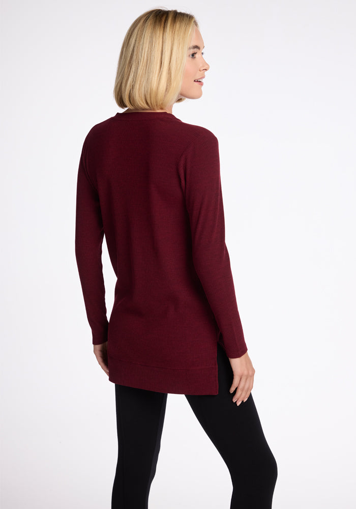 A woman with shoulder-length blonde hair is seen from the back wearing the Woolx Evie Tunic in Cranberry Melange, paired with black pants. She is smiling and slightly facing to the right against a plain white background.