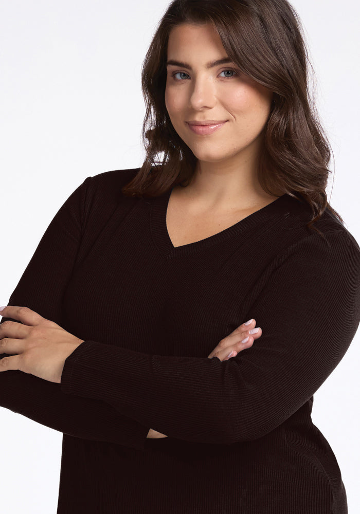 A woman with shoulder-length brown hair smiles slightly while standing with her arms crossed. She is wearing a long-sleeve, black Evie Ribbed Tunic by Woolx with a flattering V-neck fit and has a confident expression. The background is plain white.