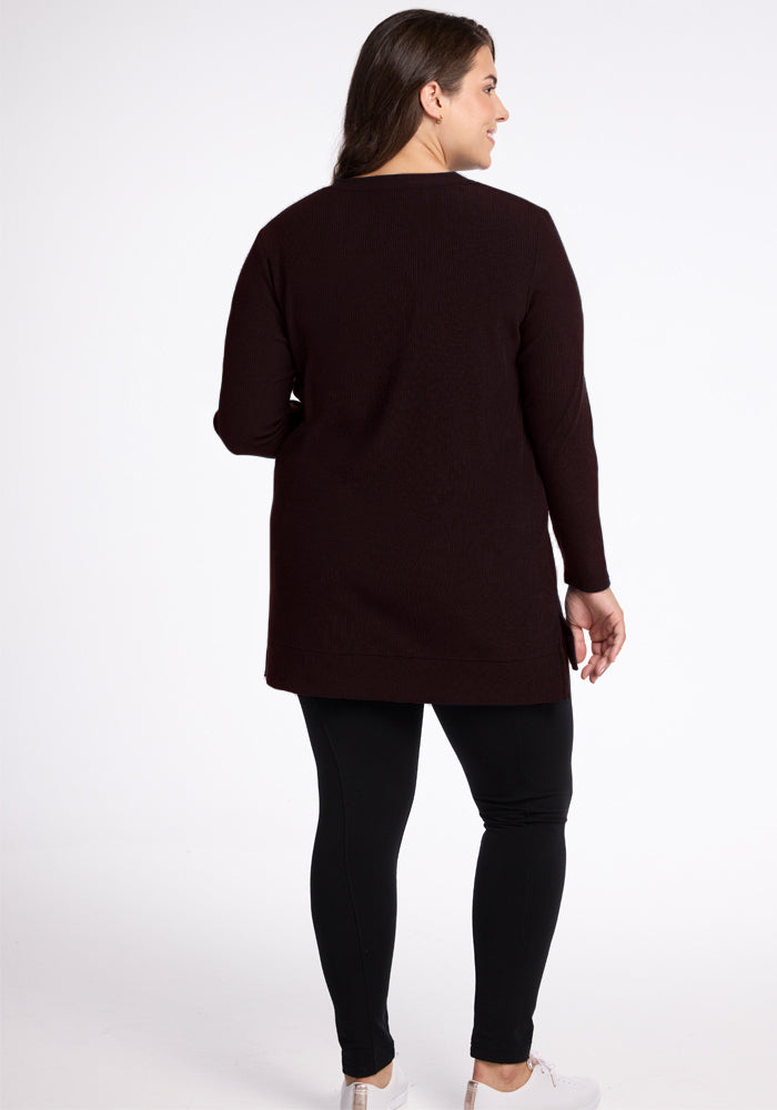 A person is standing with their back to the camera, wearing a long, dark French Roast Evie Ribbed Tunic by Woolx and black leggings. They have light-colored sneakers on and are standing against a plain, light background. The person's left hand is slightly raised, showcasing the cozy coverage of their outfit.