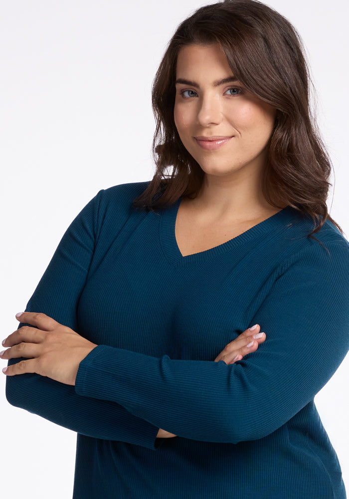 A person with long dark hair is smiling softly with arms crossed, wearing Woolx's Evie Tunic in Real Teal, which offers a flattering fit. The background is plain white.