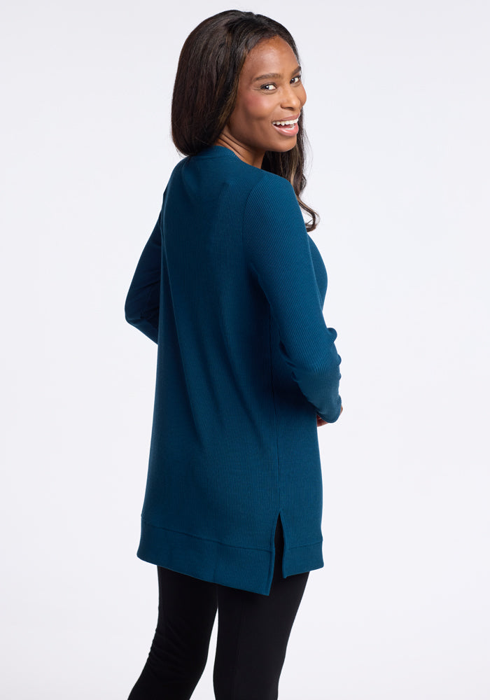 A woman with long hair, wearing the Woolx Evie Tunic in Real Teal and black pants, is smiling and looking over her shoulder against a plain white backdrop.