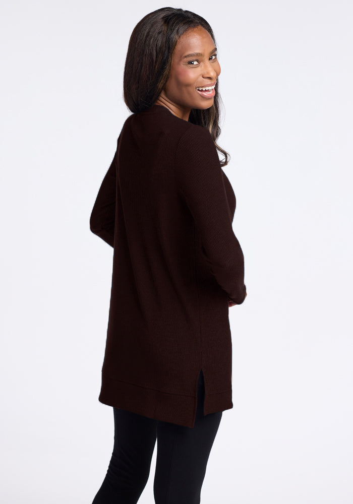 A person with long, dark hair is smiling and looking over their shoulder. They are wearing the cozy and warm Woolx Evie Ribbed Tunic in dark brown with a side slit, paired with black leggings. The background is plain white.