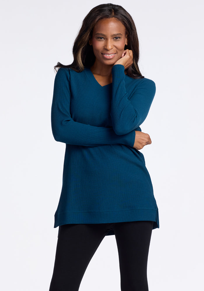 A woman poses confidently wearing the Evie Tunic in Real Teal by Woolx, paired with black leggings. Her left hand rests on her hip while her right arm is raised to her face, showcasing a slight smile on her lips. The cozy Merino wool promises comfort against the light gray background. 